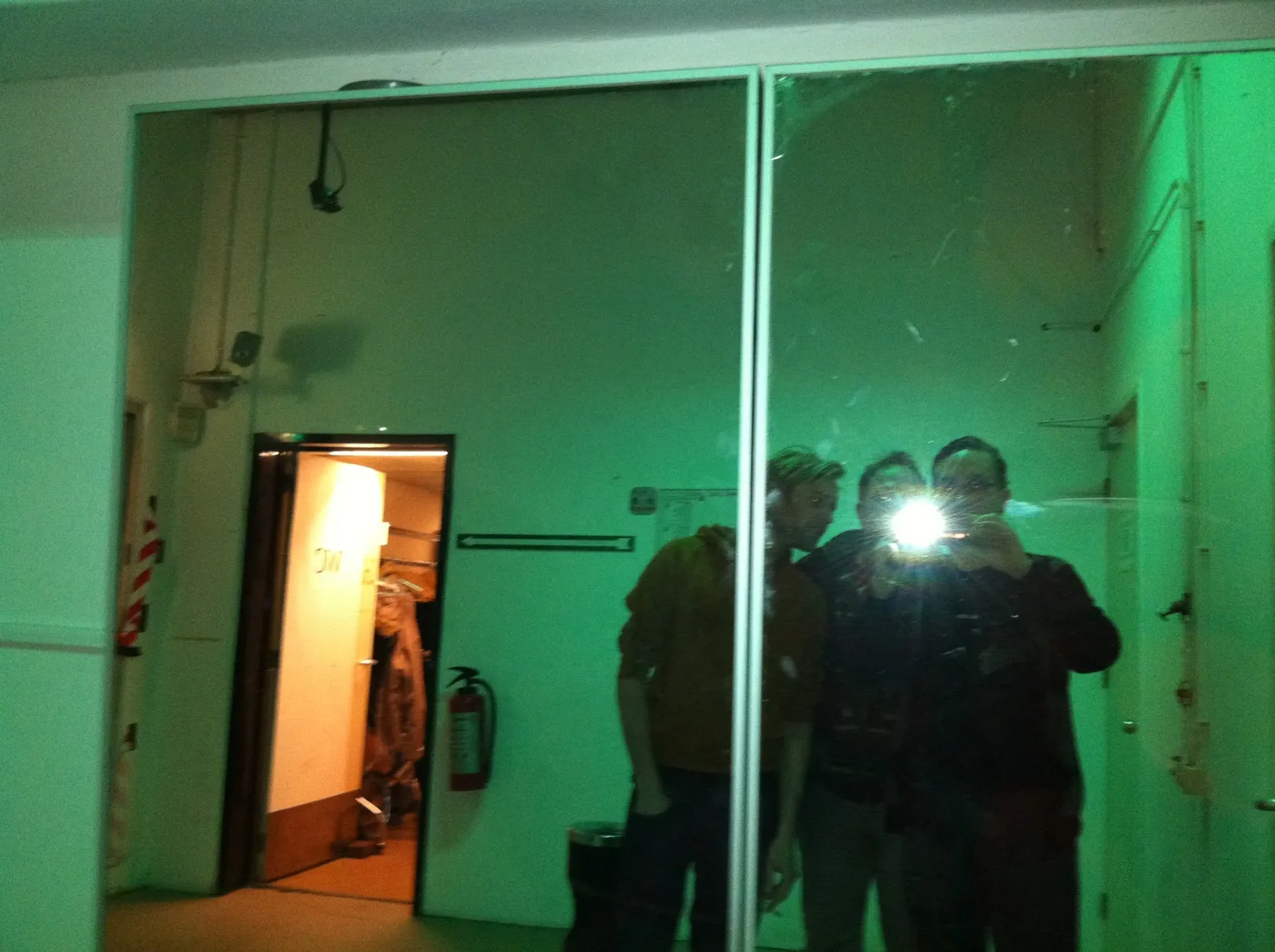 Three people in a large mirror under green light, the flash going off in the middle