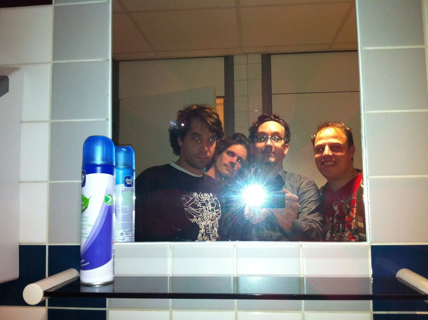 Four people squeezed into a washroom mirror, the flash in the middle, air-freshener cans on the shelf