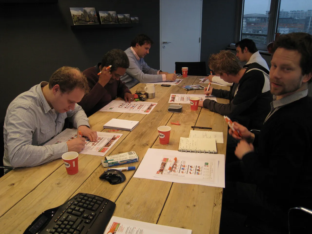 Six people at a long wooden table, participating in the workshop, marking up printed product screens with pens and markers.
