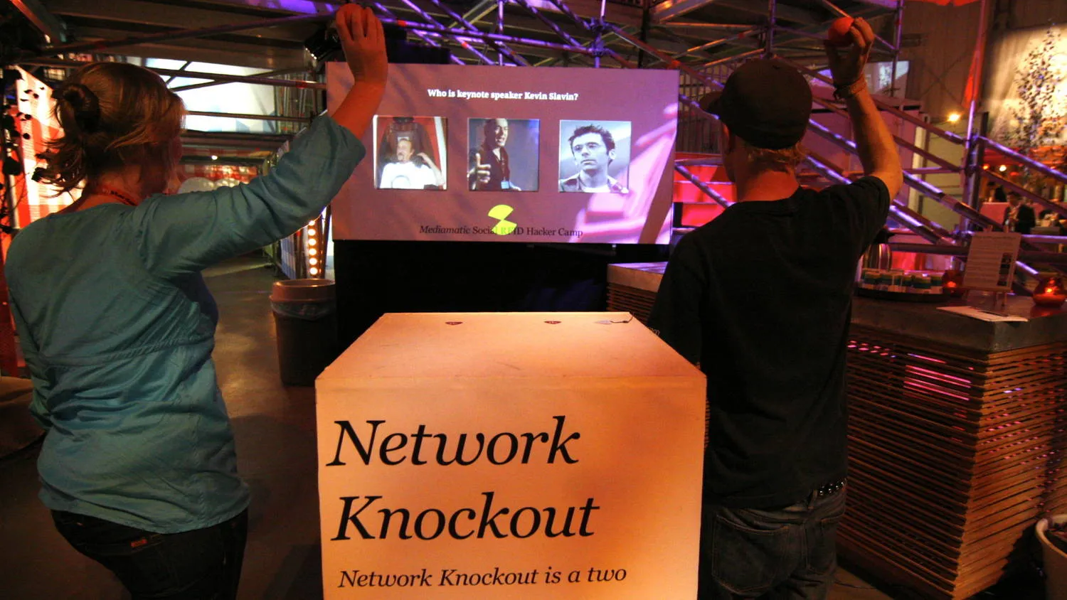 Two players, seen from behind, throwing balls at a projected quiz question with three avatars, the lit Network Knockout crate in front of them
