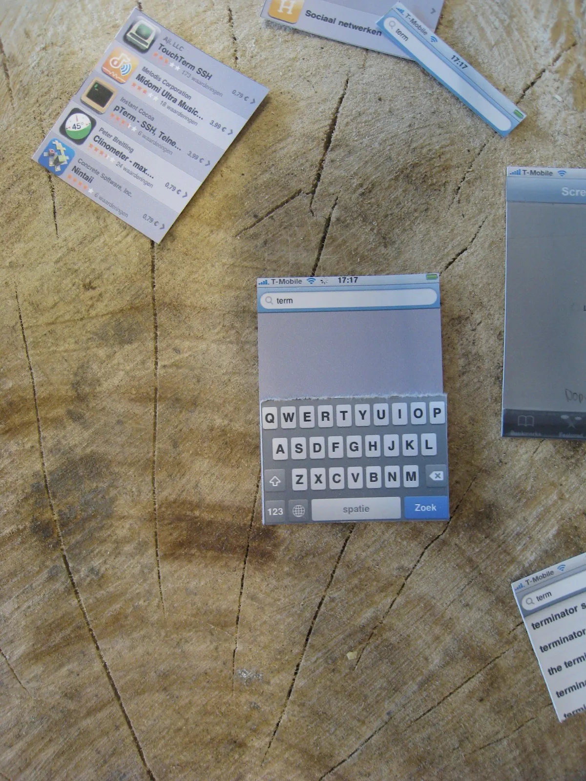 Paper prototype of search: a printed iPhone keyboard under a search field reading term, loose cut-out screens around it on a tree stump