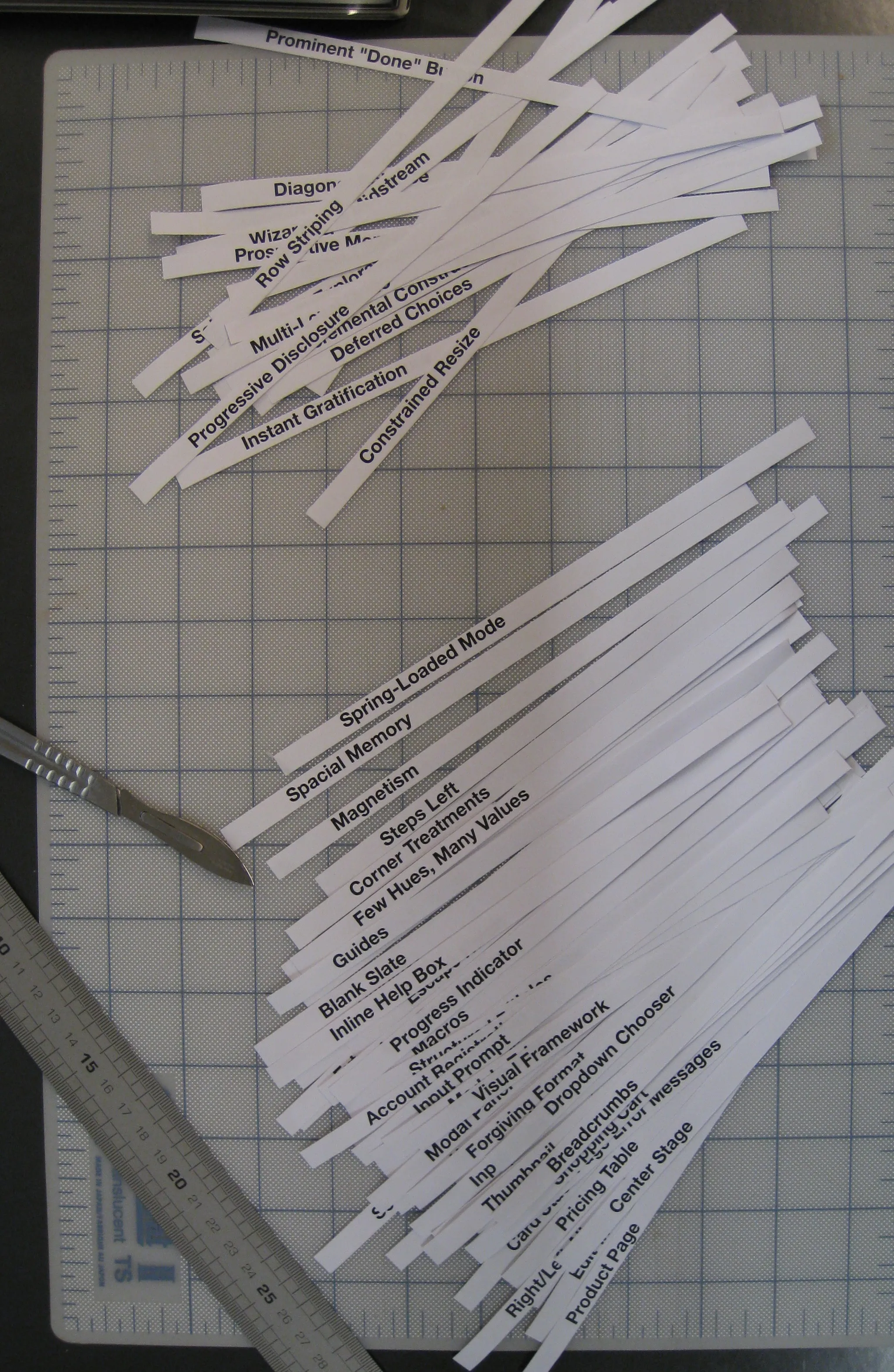 Paper strips printed with interaction design pattern names (Progressive Disclosure, Breadcrumbs, Center Stage…) sorted into piles on a cutting mat, a scalpel and a steel ruler beside them