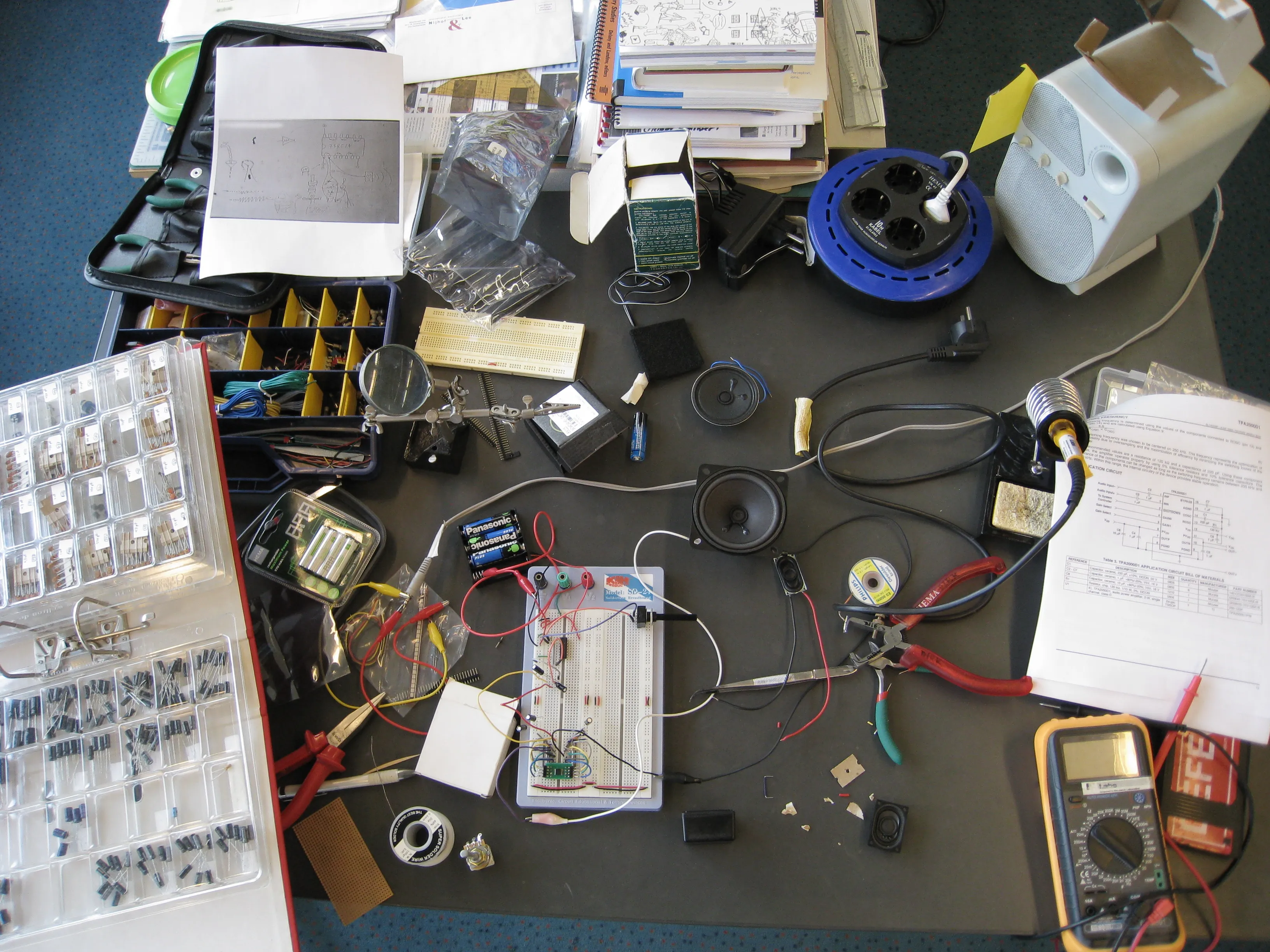 A cluttered workbench with a breadboard circuit, loose speakers, component boxes, a multimeter and datasheets
