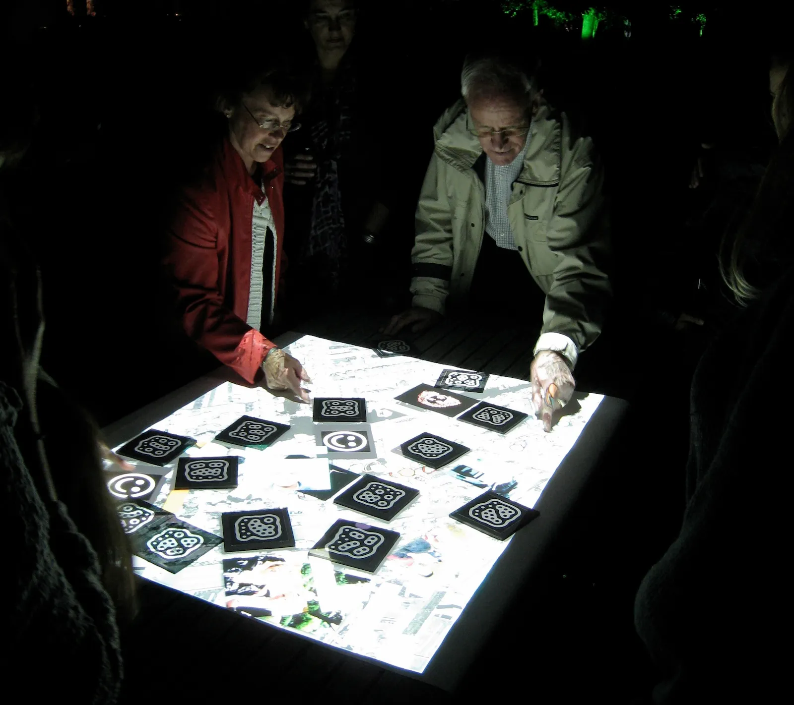 Visitors standing around the lit table at night, moving marker tiles across a projected map of their neighbourhood