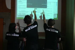 Three beta testers seen from behind, in black BETA TESTER t-shirts, pointing at a projected screen