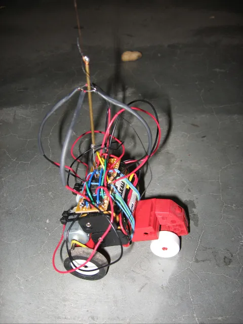 A single robot from above: a tangle of red, black and blue wires over the motor and wheels, the balloon string tied to the top of a brass rod