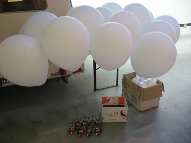 A bunch of white helium balloons tied up next to a caravan, the robots lined up on the floor below, beside the parts boxes