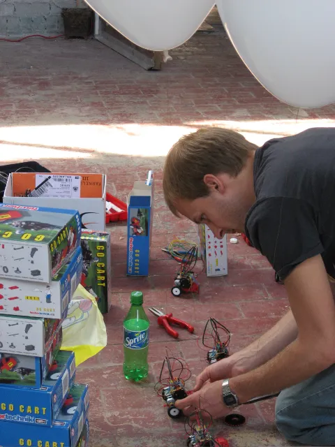 Someone kneeling on a brick floor, putting robots together between stacked Go Cart kit boxes, balloons hanging above