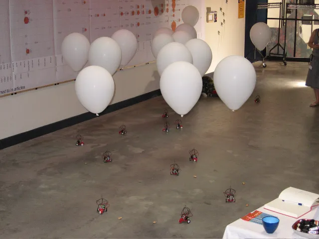The installation in a gallery with a large chart on the wall: balloons floating above the robots on a concrete floor