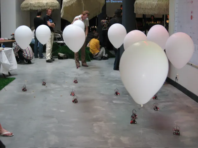 Robots and their balloons spread over a concrete floor, visitors in the space behind