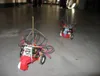 Two robots on a concrete floor, each a red toy chassis with AA batteries, a circuit board and a wire cage, the balloon string running up out of frame