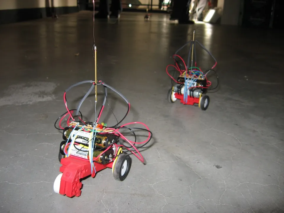 Two robots on a concrete floor, each a red toy chassis with AA batteries, a circuit board and a wire cage, the balloon string running up out of frame